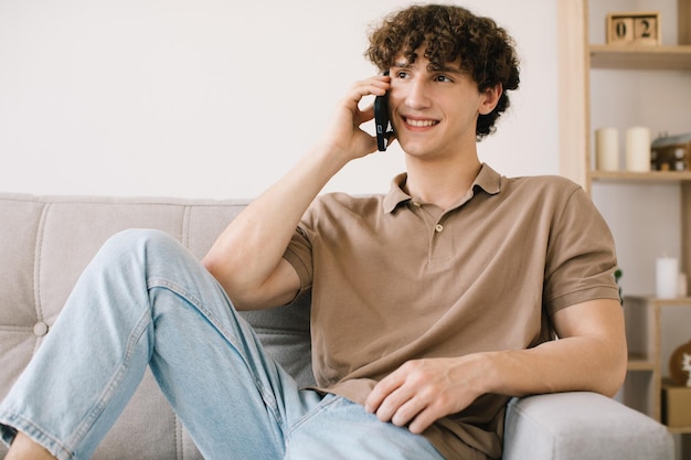 Attractive curly hair young male student talking by phone while sitting on a sofa in living room