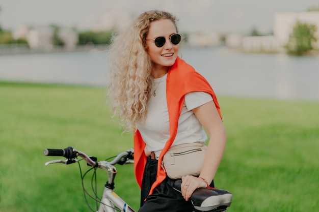 Attractive curly haired woman focused aside, wears shades, dressed in casual wear, poses on bike