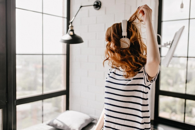 Attractive curly woman in striped tshirt dancing in her apartment Happy redhaired girl in headphones expressing positive emotions