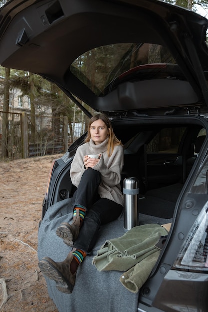 Attractive cute caucasian girl sitting in open car trunk and drinking hot tea Freedom concept