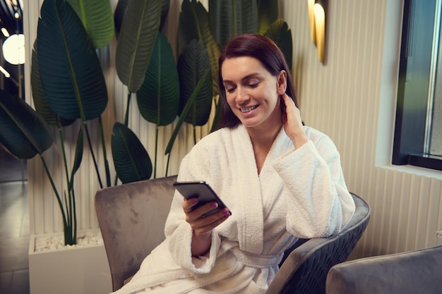 Attractive delightful Caucasian woman wearing white terry bathrobe sitting in an armchair in a luxury spa lounge zone and smiles toothy smile checking her smartphone while relaxing in wellness centre