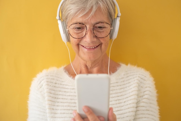 Attractive elderly woman in white sweater wearing headphones while using phone to see a video old generation and new technology