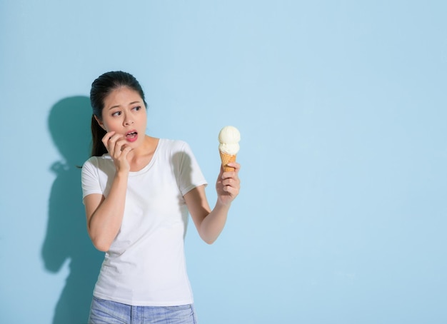 attractive elegant lady looking at fresh milk ice cream and making afraid expression because of sensitive tooth disease standing on blue wall background.