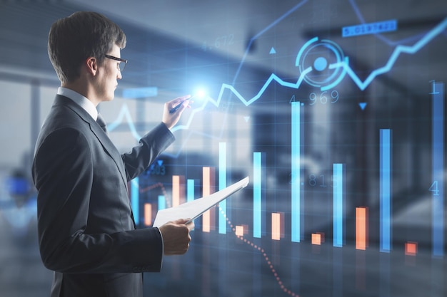 Attractive european businessman hand pointing at creative glowing forex chart on blurry office interior background Trade device financial investment and technology concept Double exposure