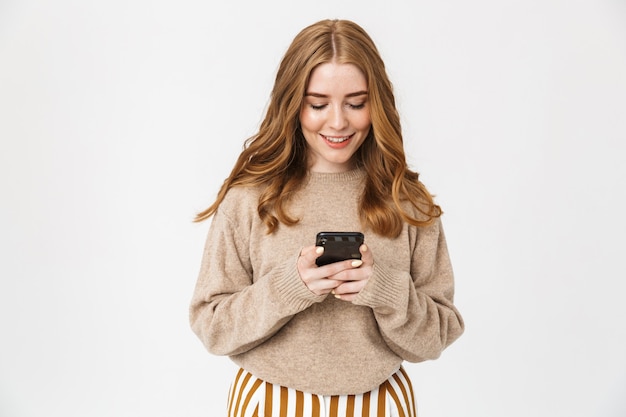 Attractive excited young girl wearing sweater standing isolated over white wall, using mobile phone