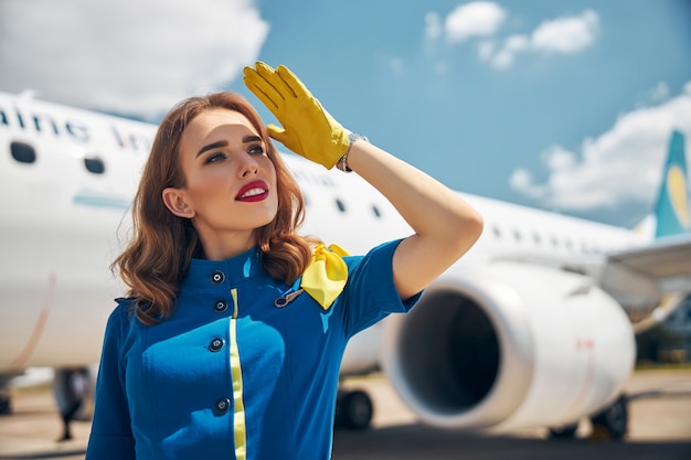 Attractive female flight attendant in yellow gloves looking away and smiling while standing outdoors with airplane on background