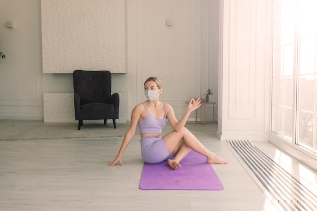 Attractive female sport fitness coach wearing white medical mask do exercises on yoga mat at a home.