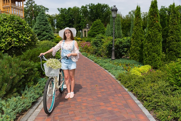 Attractive female wearing a white hat standing on footpath near her bicycle with a bouquet of little white flowers in a basket in a park with background of different trees