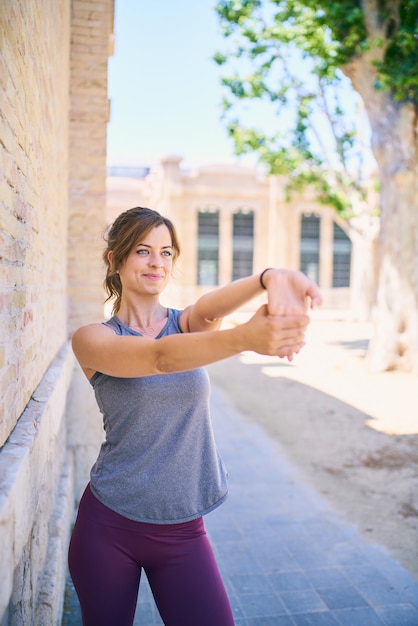 Attractive fit girl stretching at an urban location. Brunette beautiful woman wearing grey top and tight purple sportswear. Healthy lifestyle. Front shot.