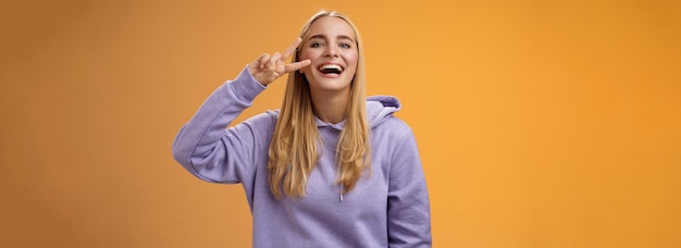 Photo attractive friendly carefree millennial blond girl in purple hoodie having fun friends laughing joyfully show peace victory hippie sign adore perfect chilly spring weather orange background