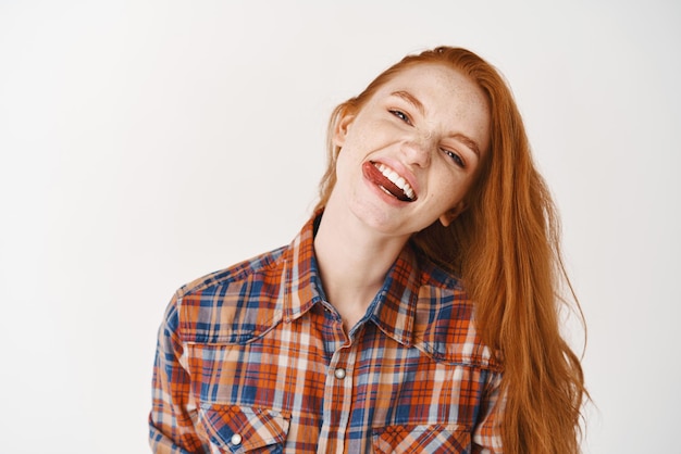 Attractive ginger girl with pale skin and freckles showing tongue having fun and looking happy standing on white background