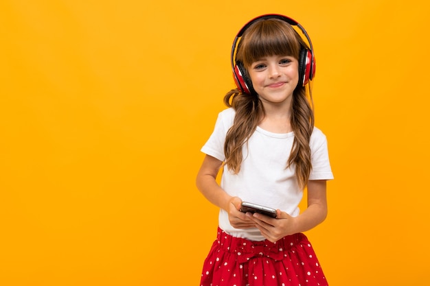 Attractive girl enjoys listening to music on headphones and holding a phone in her hands on an orange studio background.