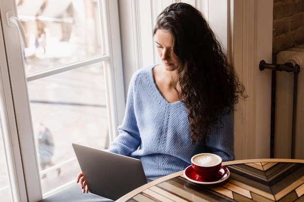 Attractive girl freelancer is working online on laptop and drinking coffee in cafe Confident business woman manager with laptop is working with colleagues online in cafe