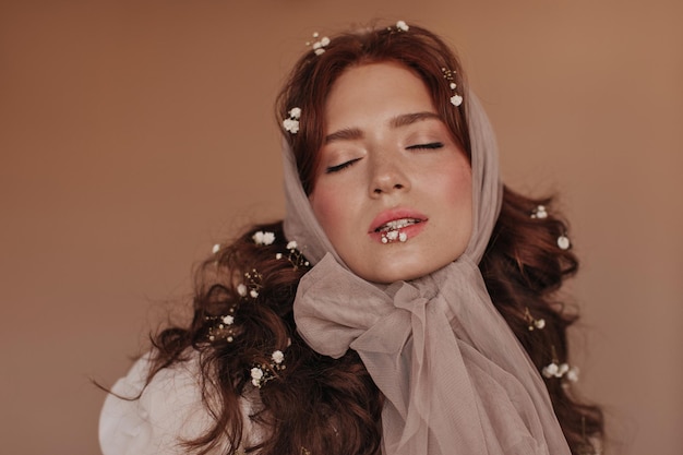 Attractive girl holding small white flower in mouth Lady in beige headscarp posing with closed eyes