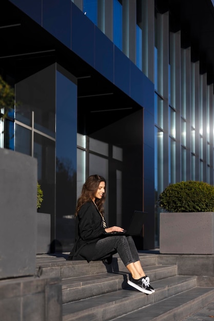 Attractive girl is working on a laptop squatting on the stairs near a modern glass office Modern young girl freelancer work online education online