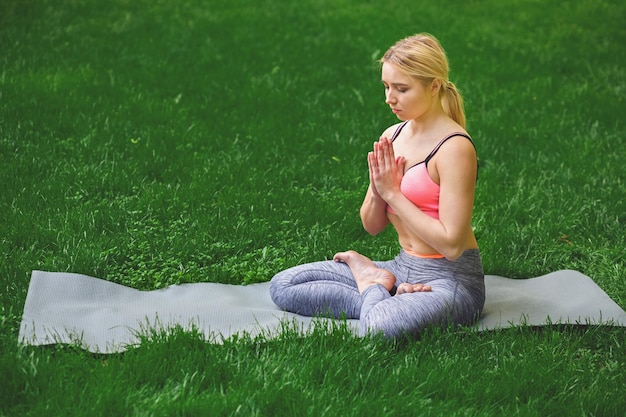 Attractive girl practicing yoga sitting in padmasana. Young woman in lotus pose, namaste with closed eyes on mat outdoors, copy space. Wellness, calmness, relax, healthy, active lifestyle concept