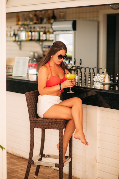 Attractive girl resting on the beach bar, drink a refreshing cocktail. The bright bathing suits and sunglasses.