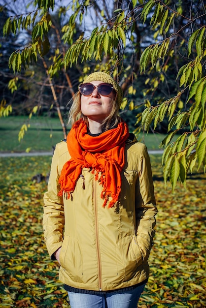 Attractive girl in sunglasses and red scarf smiling on the background of yellow foliage in autumn park. Young positive woman enjoying walk on fall sunny day.