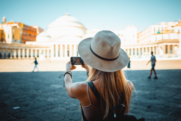 Attractive Girl tourist in hat with backpack exploring new city in Europe at summer and using her phone to take photo