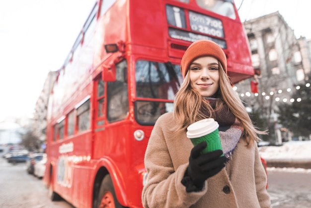 attractive girl in warm spring clothes,standing with a paper cup of coffee in the hands of a red tourist bus,watching the camera and smiling