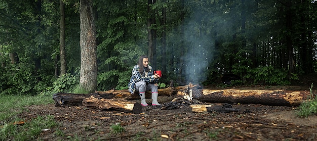 An attractive girl with a cup in her hand sits on a log and warms herself near a fire in the forest.