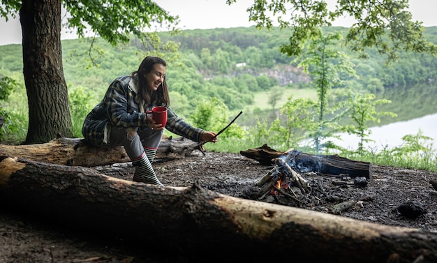 An attractive girl with a cup in her hand sits on a log and warms herself near a fire in the forest.