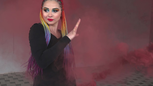 Attractive girl with multicolored braids and makeup in an black dress. Posing hiding behind red artificial smoke against the background of an building in a spring city.