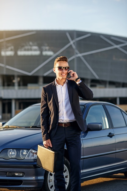 Attractive handsome businessman with diplomat talking on smartphone near the car