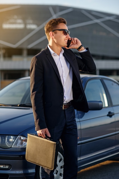 Attractive handsome businessman with diplomat talking on smartphone near the car