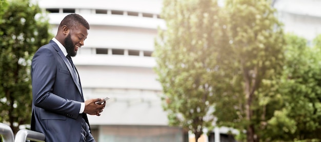 Attractive happy businessman is texting on cell phone near modern office building side view with