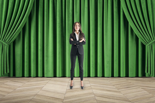 Attractive happy businesswoman with folded arms standing in front of green curtain and wooden flooring Cinema stage and theater