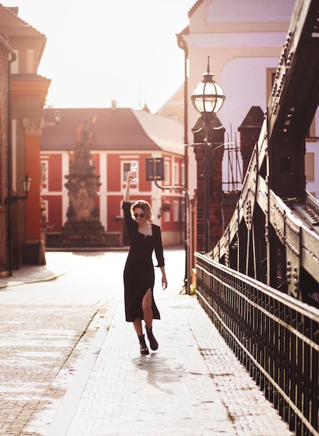 Attractive happy girl in a black dress walks along a city street with a glass of wine Glamorous lifestyle Fashion