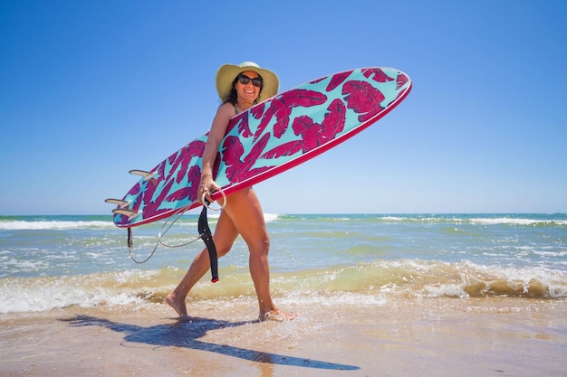 Attractive happy middleaged woman wearing a hat sunglasses and carrying a surfboard on the beach