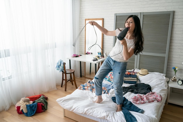 attractive happy young asian woman in casual wear singing at hair dryer on bed in home cozy bedroom. tourism business trip people and luggage concept. lady packing travel bag relax having fun alone