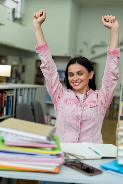 Attractive happy young girl student studying at the college library sitting at the desk writing and reading stretching hands