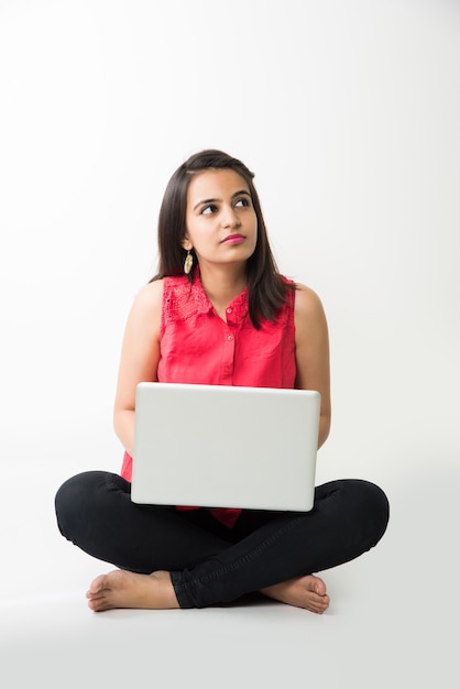 Attractive Indian Asian girl student studying on laptop computer with pile of books, sitting isolated over white floor