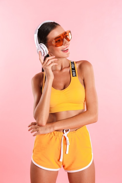 Attractive joyful young slim brunette woman dressed in sportswear standing isolated over pink wall, listening to music with headphones