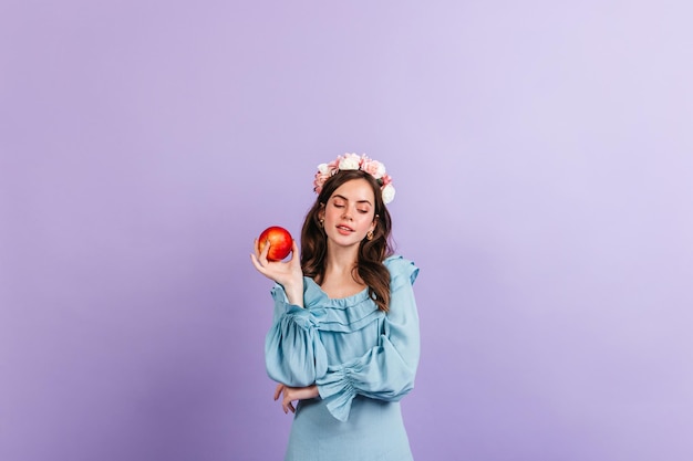 Attractive lady modestly looks down holding large apple on purple background Girl with curly hair posing in blue dress