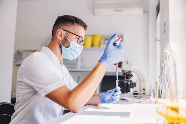 Attractive male lab assistant with rubber gloves and face mask holding test tube with blood and doing research for the cure for corona virus.