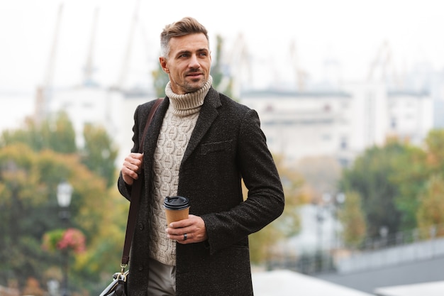 Attractive man wearing jacket walking outdoors, holding cup of takeaway coffee