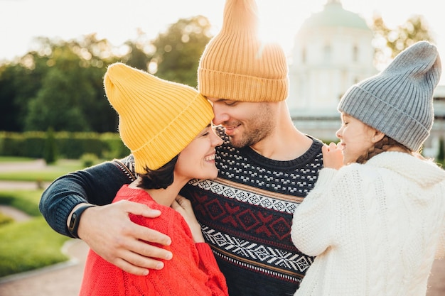 Attractive man wears yellow warm hat embraces his wife and daughter looks at them with great love Adorable little girl feels support from parents smiles happily while being on fathers hands