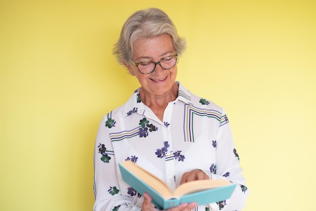 Attractive mature caucasian woman holding a book in hand enjoying reading isolated on yellow background