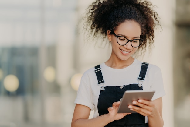 Attractive millennial girl with crisp hair, holds digital tablet, surfes social networks, wears optical glasses