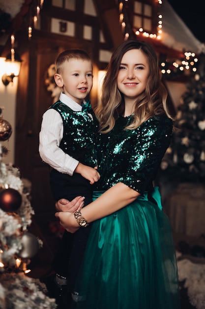 Attractive mother with long wavy hair in green coctail dress holds her little caucasian son on her arms and smiles