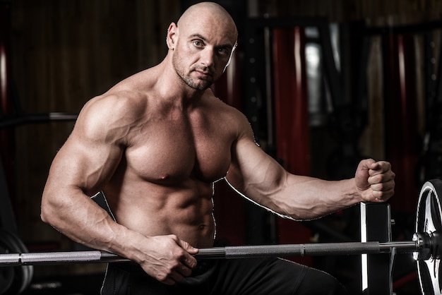 Attractive muscular bodybuilder guy prepare to do exercises with barbell in a gym. Guy bodybuilder tired in gym. Young Man Bodybuilder Resting In Gym After Exercising