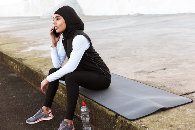 Photo attractive muslim sportswoman wearing hijab outdoors, sitting on a fitness mat, taking on mobile phone