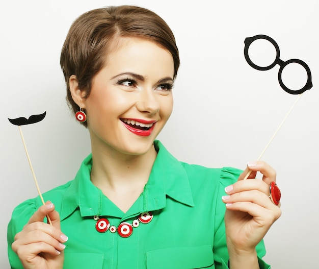 Attractive playful young woman holding mustache and glasses on a stick. Ready for party.