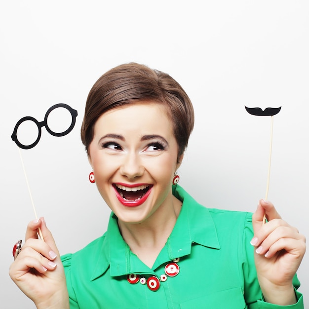 Attractive playful young woman holding mustache and glasses on a stick. Ready for party.