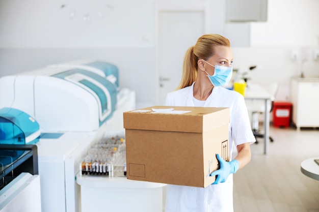 Attractive positive blond lab assistant carrying box with vaccines for Covid 19. Laboratory interior.