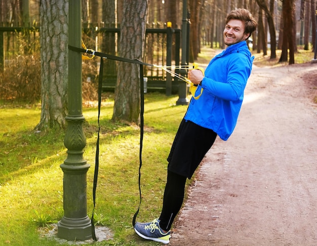 Attractive positive male in a blue raincoat exercising in a park with trx fitness strips.